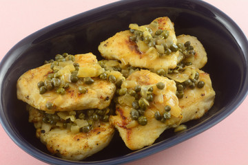 Closeup of cooked chicken breast pieces with caper gravy sauce in blue serving bowl on pink background
