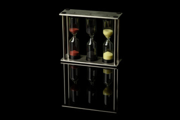 A set of three hourglasses with colored sand stands on a black glossy glass, in which their reflection is visible.