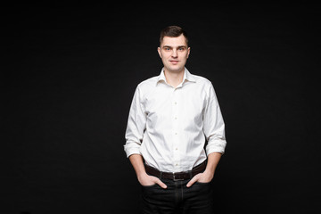 Waist up of handsome young man in white shirt holding hands in pockets while posing against black...