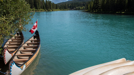Canoe boat docked by the shore. Turquoise water from the Bow River and Canada Flag background