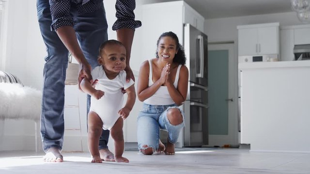 Parents Encouraging Smiling Baby Son To Take First Steps And Walk At Home