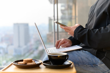 casual wearing man operating smartphone and laptop with coffee and cookies nearby on the balcony concept of work from home
