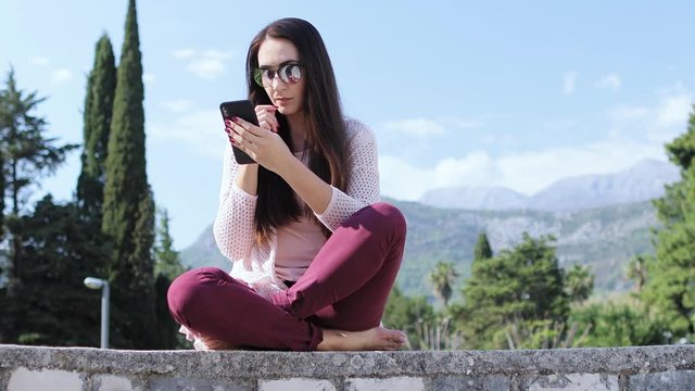 woman texting and chatting on phone outdoors sitting in park with mountains view. Long hair bunette woman texting and using phone outdoors. 