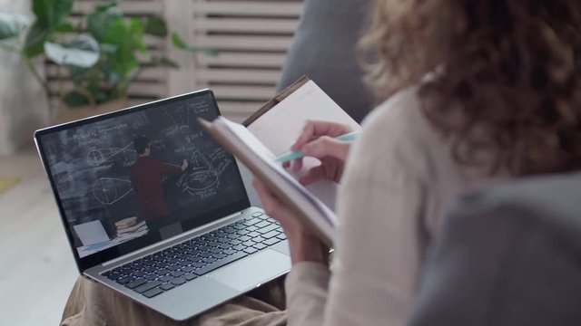 Rear view of unrecognizable curly-haired woman sitting on couch at laptop, having online lesson and making notes in notebook