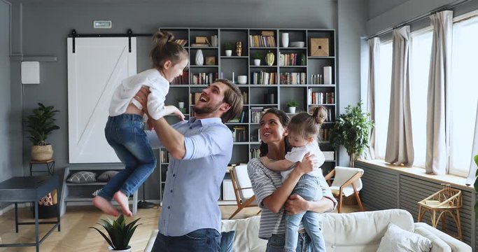 Young adult parents mum and dad playing active game with small adorable children lifting throwing little cute daughters up in living room. Happy carefree family having fun enjoying lifestyle at home.