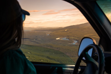 Woman with sunglasses & hat driving a car with Yukon scenic background in summer time. Summer Canadian view. 