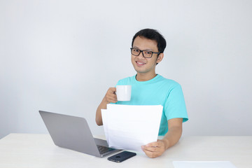 Young Asian Man is smile and happy when working on a laptop and document with white mug on hand. Indonesian man wearing blue shirt.