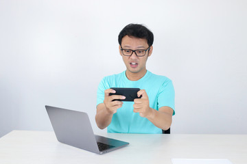 Wow face of Young Asian man shocked what he see in phone with laptop beside it. Indonesian man wearing blue shirt.