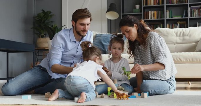 Happy relaxed family young adult parents and cute small kids daughters play toys sit on warm floor carpet. Loving mother, father have fun with two little children enjoy free time games in living room.