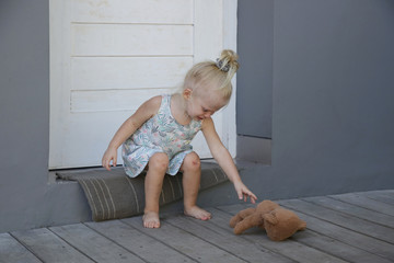 Little girl sitting outside alone trying to reach for her teddy bear. Concept of abused naglected child.