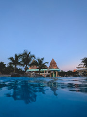 Fototapeta na wymiar Amazing pool with building with straw roof, palms, and the sunset blue sky, Mexico