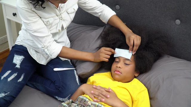 4K African American mother put fever patch on daughter head and using digital oral thermometer checking the temperature of illness with cold and flu girl. Motherhood and children healthcare concept.