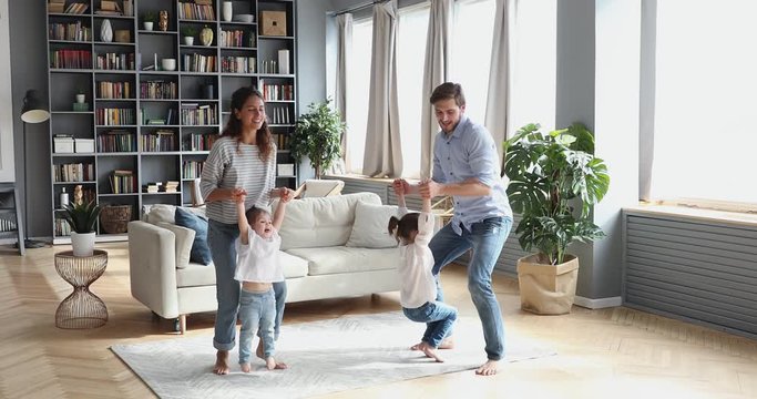 Happy active young parents dance with cute little kids daughters in modern living room interior. Adult couple having fun lifting children up enjoy family life funny weekend activity in cozy apartment.