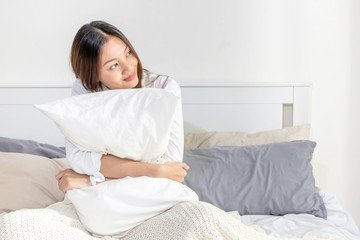 Portrait of smiling relax young beautiful asian woman sitting on the bed with hugging white pillow in bedroom. Happy pretty girl have a good sleep at night and wake up in the morning with freshness.