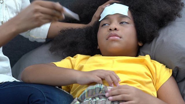 4K African American mother put fever patch on daughter head and using digital oral thermometer checking the temperature of illness with cold and flu girl. Motherhood and children healthcare concept.