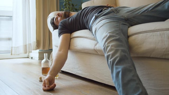 Drunk man sleeping on the couch dressed. Nearby is an unfinished bottle of whiskey and a glass. Young debauchee in his apartment after party