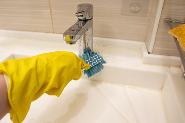 A hand in a yellow glove cleans the sink with a blue brush on the handle. The concept of homework, disinfection