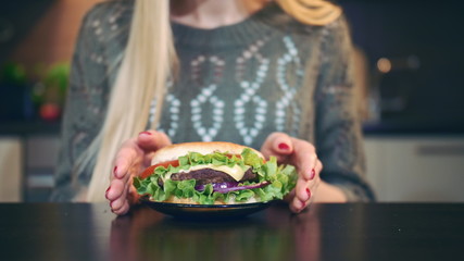Crop view of Young lady preferring hamburger to salad. Attractive young woman choosing to eat healthy hamburger for breakfast while sitting at table in stylish kitchen.