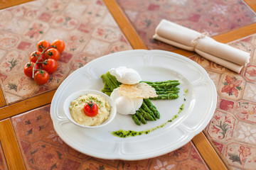 poached egg with grilled asparagus sprouts, served on a white oval plate under a cheese crust with mashed potatoes and tomatoes. vintage dining table and fabric napkin. top view