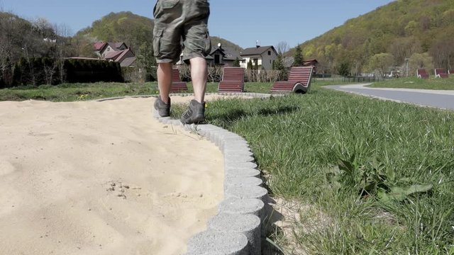 Man walking along a public sandbox has eventually sat and rested on a public wooden chair. No people. COVID 19.