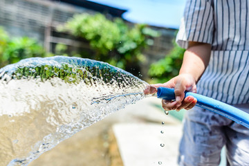 The boy is holding a hose to spray water on the tree. He tried to use his little finger to cover the pipe a little to increase water pressure. What happened makes him enjoy the watering of plants.