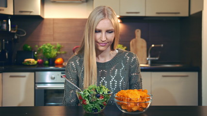 Young lady preferring salad to crisp. Beautiful young woman choosing to eat healthy crisp for supper while sitting at table in stylish kitchen and she eats these salad.