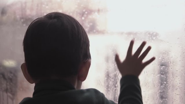 Slow motion video of a sad kid playing with condensation on the glass window on a rainy day during virus quarantine  at home