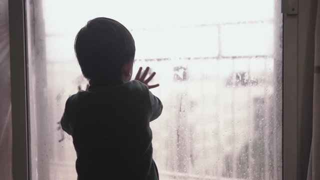 Cute little kid asian boy putting his hands on the glass of a window covered in condensation on a rainy day during lockdown missing freedom and being in the street
