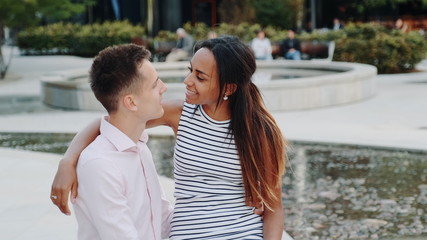 Fototapeta premium Close-up of cheerful multiracial couple having date in a beautiful park. They sitting on the bench, hugging, talking and looking at each other.
