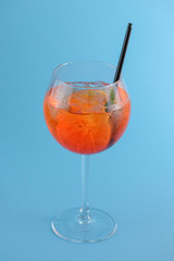 Cold summer alcohol cocktail with ice and orange in a wine glass and black plastic straw on light pastel blue background
