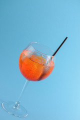Cold summer alcohol cocktail with ice and orange in a wine glass and black plastic straw on light pastel blue background