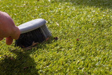Man brushing rubbish on artifiicial grass turf with a hand brush in sunshine.  Garden chore concept