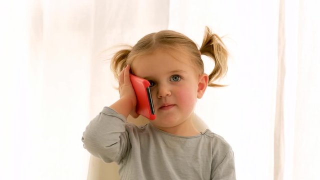 Little girl with ponytails pretending to speak on smartphone while sitting on chair and playing on weekend day at home