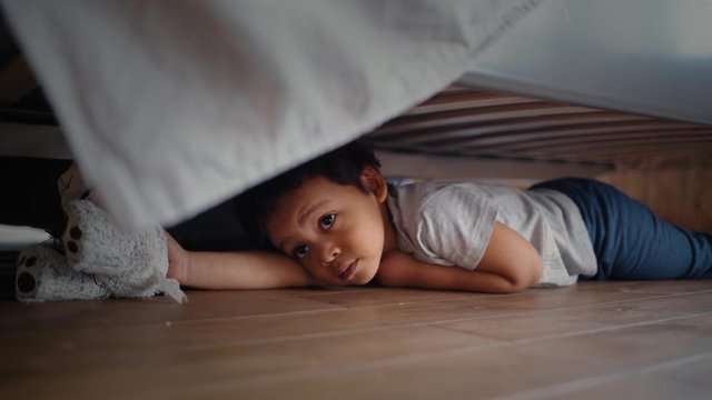 Hiding baby lies laughing and crawling out from under the bed. Child looking for something under the bed.