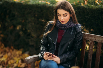 Stylish beautiful lady in a black leather jacket with a black bag and burgundy blouse. Attractive young woman use phone