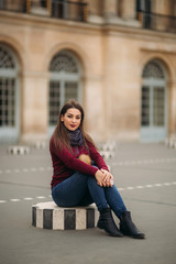 Fototapeta premium Attractive young womanl in burgundy blouse with a gray scarf sits on a striped dais. Happy woman in Paris