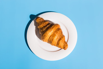 Bakery products baked croissant on white plate. Blue background, top view close-up. Pop art style. Summer shadows. Delicious and food concept.