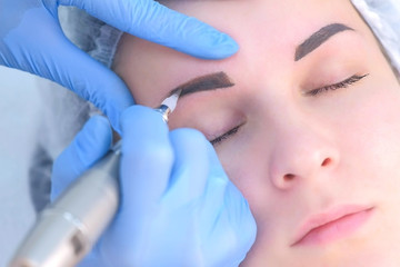 Cosmetologist making eyebrows microblading procedure in beauty salon for girl using tattoo apparatus, face closeup. Beautician in gloves is doing permanent eyebrow makeup. Beauty industry concept.