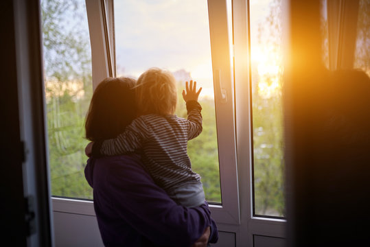 Mother with baby looks out the window at sunset in isolation at home for virus outbreak. Stay home concept