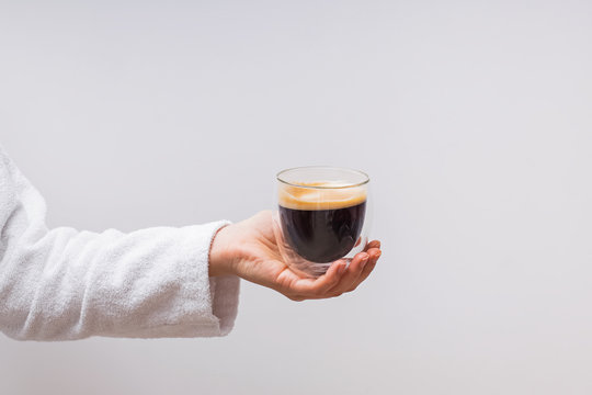 Woman's hand in white bathrobe holding glass double wall cup with espresso coffee