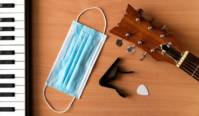 Piano keys, acoustic guitar, capo, pick and blue medical mask lie on a wooden background. Coronavirus epidemic music concept