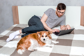Young man working on a laptop in bed at home and dog beside. Concept Working remotely