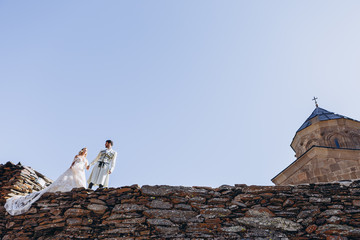 Fototapeta na wymiar stylish Georgian groom dressed in a national costume leads his bride holding her hand on a rocky cliff against the backdrop of the sky and the church where the wedding was held