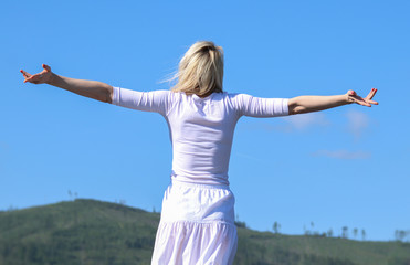 Fototapeta na wymiar Happy young woman in a white dress spreading her hands in the background of the green hills and the blue sky