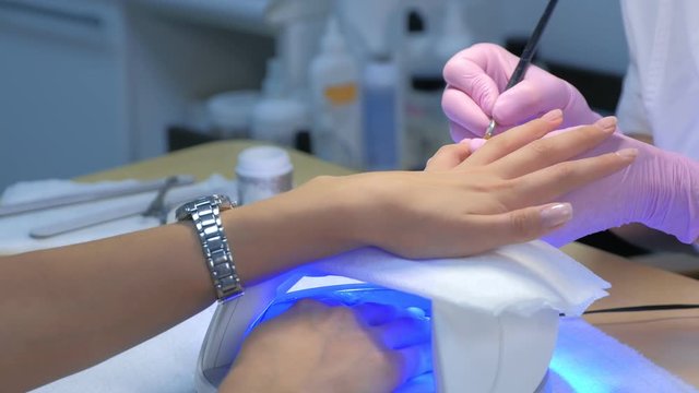 Manicurist master is painting client's nails gel shellac, another hand is drying in UV lamp in beauty salon, hands closeup. Manicurist in gloves is making manicure.
