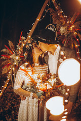 Stylish couple of brides, pretty woman in wedding dress and young man in fedora hat. Romantic wedding ceremony in nature. The lights of the electric garland illuminate the wedding party.