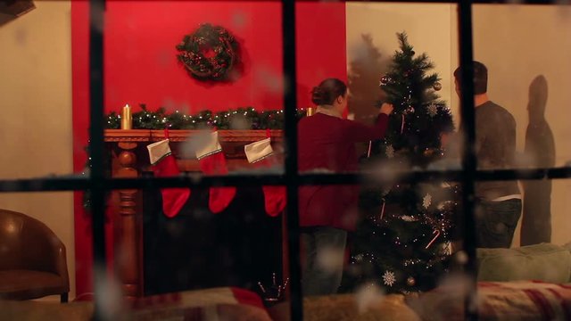 Couple at home decorating the Christmas Tree with snow falling outside. Looking in through the window. Stock Video Clip Footage