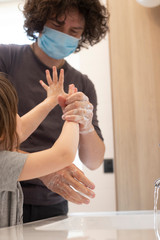 Caucasian father wearing face mask to avoid contagion teaching little child girl how to wash hands in bathroom during covid-19 pandemic lockdown. Dad and baby daughter washing hands at sink. Vertical.