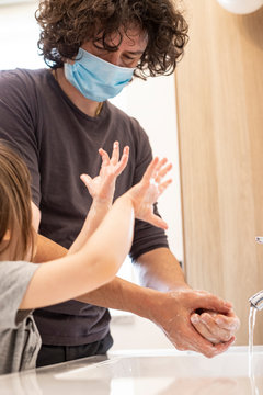 Caucasian Father Wearing Face Mask To Avoid Contagion Teaching Little Child Girl How To Wash Hands In Bathroom During Covid-19 Pandemic Lockdown. Dad And Baby Daughter Washing Hands At Sink. Vertical.