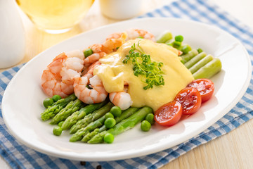 Hot shrimp with fresh vegetables and a side dish of green asparagus, watered with hollandaise sauce and garnished with fresh microgreens.
